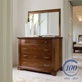 Comò classico in legno ciliegio con specchiera coordinata, parte della collezione Arte Piombini. Il mobile ha quattro cassetti con maniglie in ottone finemente lavorate e intarsi decorativi sui frontali. Sopra il comò, specchiato da una grande finestra, si trovano eleganti vasi bianchi e alcuni libri. Nell’angolo in basso a destra, il logo del centenario (1925–2025) con la scritta “100 anni” e una corona d’alloro. Sullo sfondo si intravede una camera da letto arredata nello stesso stile.