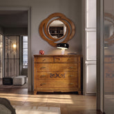 Interior di una lussuosa e sofisticata zona notte con una grande apertura che conduce a una cabina armadio, illuminata dalla luce solare calda che entra da destra. Al centro, su un pavimento in parquet a lisca di pesce, è posizionato il Como Classico Cinque Cassetti con Segreto della collezione Arte d'Este di Piombini, in finitura ciliegioSopra il comò, si trovano la lampada Snoopy di Flos, un fermacarte elefantino di Hermes rosso lucido e una Specchiera Classica Ovale sempre della collezione D'Este.