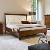 Letto matrimoniale classico in legno di ciliegio con testata imbottita in vera pelle color ecrù, decorata con raffinata lavorazione capitonné. Il design elegante è accompagnato da comodini coordinati e da un comò con intarsi, tutti parte della collezione Arte Piombini. La stanza, luminosa e accogliente, è caratterizzata da ampie finestre e arredi in stile tradizionale. In basso a destra è presente il logo del centenario Piombini (1925–2025) con la scritta “100 anni” racchiusa in una corona d’alloro