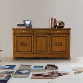 Credenza classica D'Este P3003 in legno, con tre porte decorate, tre cassetti e finitura calda effetto ciliegio, ambientata in un elegante soggiorno.