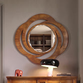Una specchiera ovale con una cornice in legno intagliato con motivi a riccioli e volute traforate, con finitura color ciliegio. Al centro c'è uno specchio circolare. La specchiera è appesa su una parete sopra un comò in legno, con una lampada moderna, libri e un piccolo elefante rosso sulla superficie.