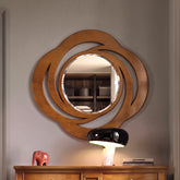 Una specchiera ovale con una cornice in legno intagliato con motivi a riccioli e volute traforate, con finitura color ciliegio. Al centro c'è uno specchio circolare. La specchiera è appesa su una parete sopra un comò in legno, con una lampada moderna, libri e un piccolo elefante rosso sulla superficie.