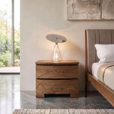 Inquadratura frontale simmetrica del comodino contenitore moderno a due cassetti in legno di noce, codice SKU 8271D, con linee sagomate e piedini squadrati, sormontato da una lampada da tavolo accesa vicino al letto.