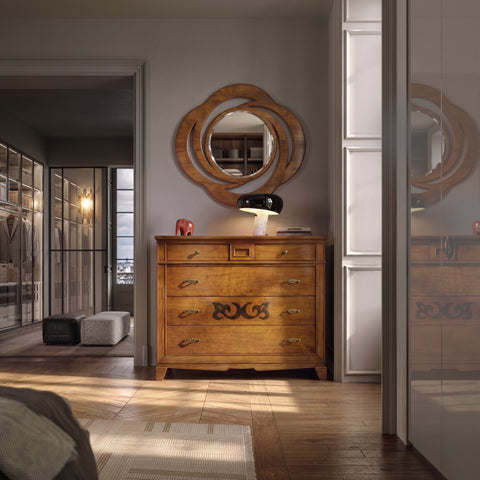 Interior di una lussuosa e sofisticata zona notte con una grande apertura che conduce a una cabina armadio, illuminata dalla luce solare calda che entra da destra. Al centro, su un pavimento in parquet a lisca di pesce, è posizionato il Como Classico Cinque Cassetti con Segreto della collezione Arte d'Este di Piombini, in finitura ciliegioSopra il comò, si trovano la lampada Snoopy di Flos, un fermacarte elefantino di Hermes rosso lucido e una Specchiera Classica Ovale sempre della collezione D'Este.
