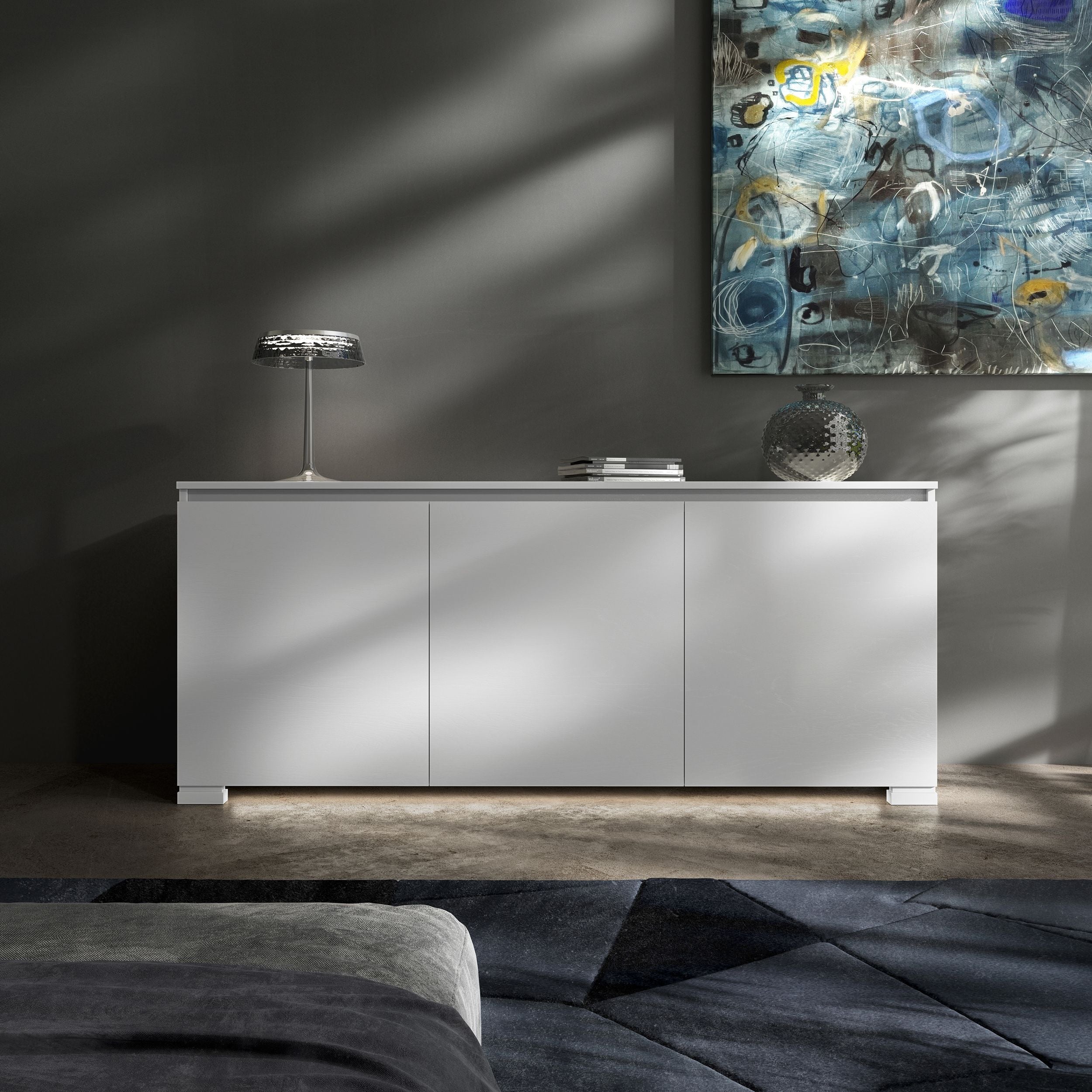 Credenza Modigliani Piombini a 3 ante in finitura Bianco Laccato con anta liscia; ambientazione in stile Soft Modern con lampada Flos Bon Jour, vaso Venini Monofiore Balloton fumè, tappeto geometrico blu e divano moderno grigio.