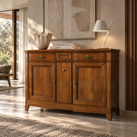 La credenza D’Este Piombini, vista di tre quarti, mostra il legno dai toni ambrati, le due ante incorniciate e i tre cassetti. Sul piano, un vaso dai bordi ondulati, libri e una lampada chiara. La luce del giardino attraversa le tende e accarezza il mobile, in un soggiorno dall’eleganza raccolta.