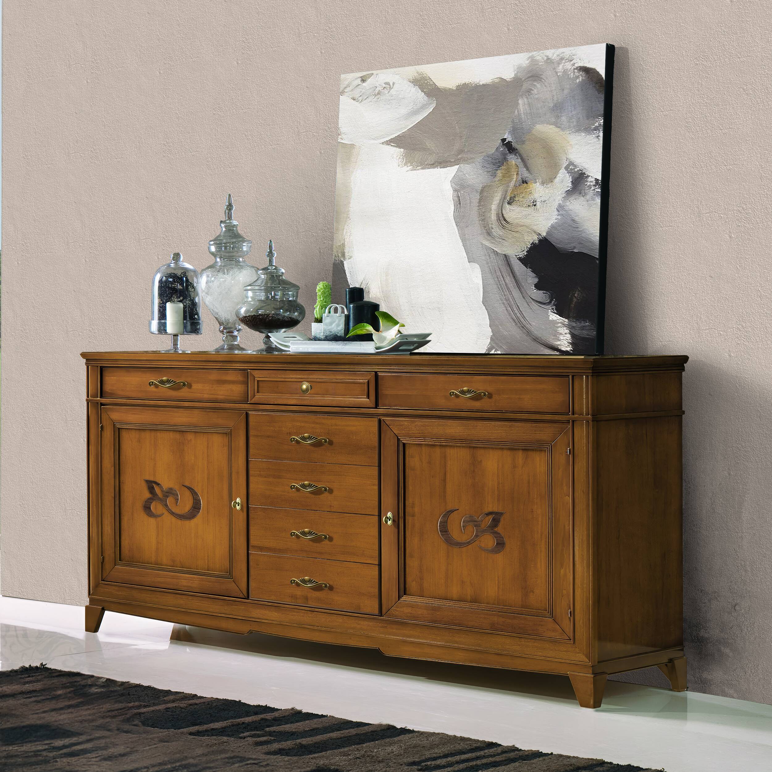 Vista angolata della credenza modello P3010 della collezione Arte d'Este di Piombini, in finitura GRN14 Bassano chiaro anticato. Mobile classico italiano in legno massello, con due ante laterali decorate e cinque cassetti centrali, arricchito da eleganti maniglie dorate. Sopra, un quadro astratto e contenitori in vetro decorativi.