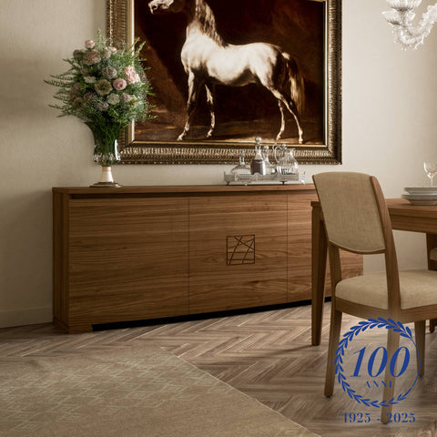 Credenza moderna a tre porte in legno di noce della collezione Modigliani, con intarsio centrale e piano superiore decorato. Ambientazione elegante con tavolo coordinato e dipinto di cavallo sullo sfondo. Logo del centenario 1925–2025 in basso a destra.