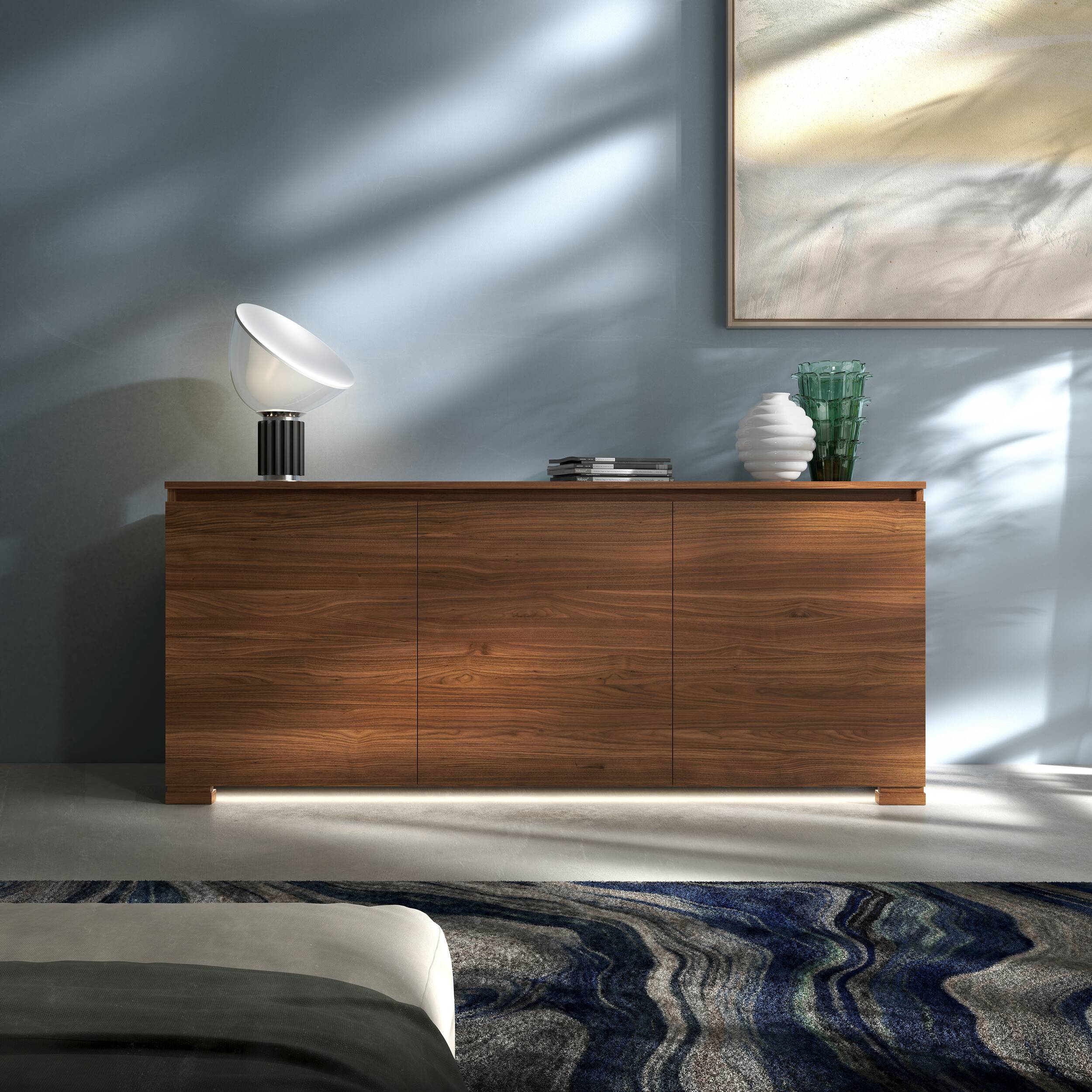 Credenza Modigliani Piombini a 3 ante in finitura Noce Naturale con Ante Lisce; ambientazione in stile Contemporaneo Moderno con lampada Flos Taccia, vaso Venini Deco bianco e vaso Venini Ritaglio verde, tappeto contemporaneo blu e divano moderno grigio.