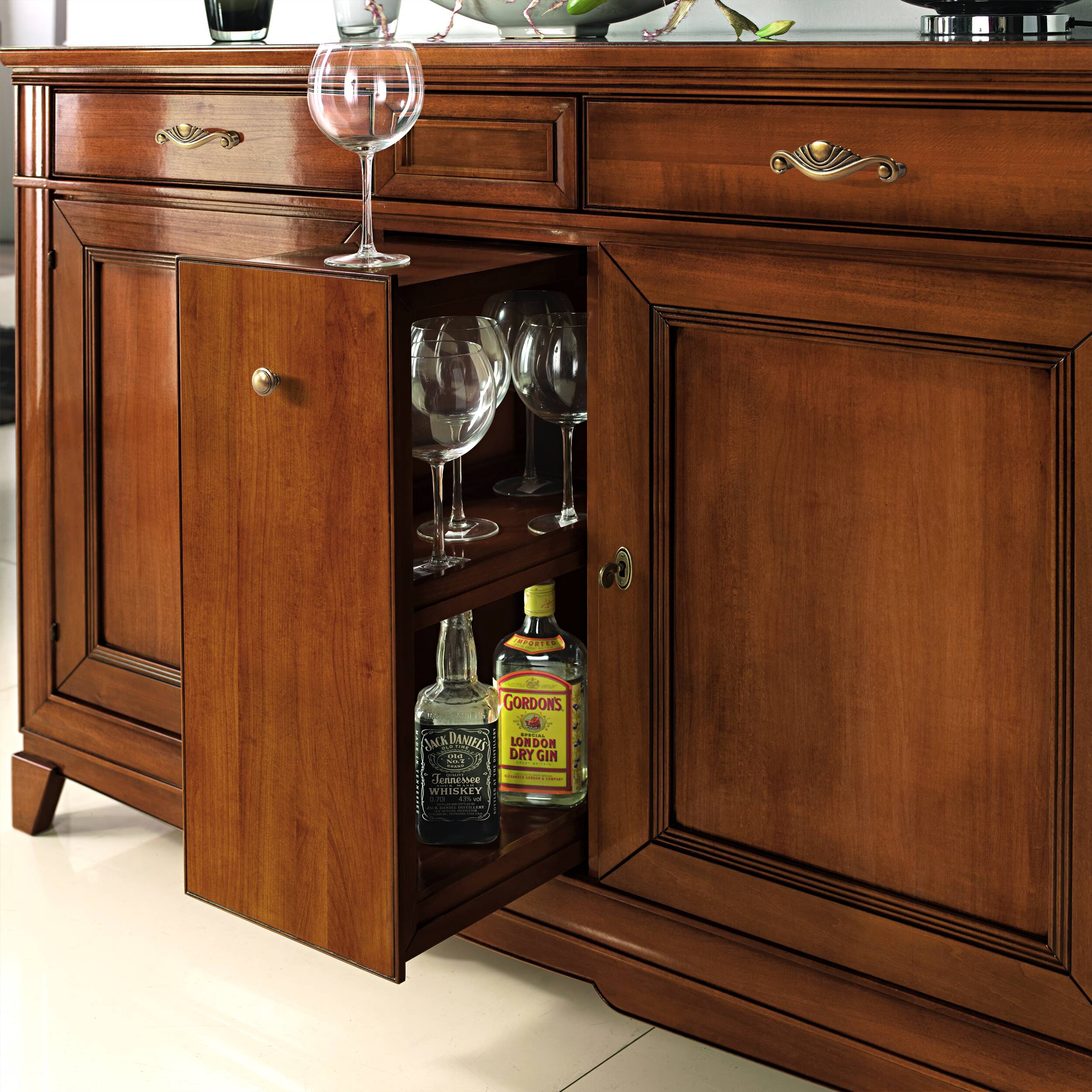 Dettaglio della credenza classica in legno di ciliegio con anta centrale portabottiglie aperta, dotata di ripiani interni per bottiglie e bicchieri. Il mobile presenta due ante laterali, eleganti maniglie in ottone e una finitura calda. Parte della collezione Arte d’Este Piombini.