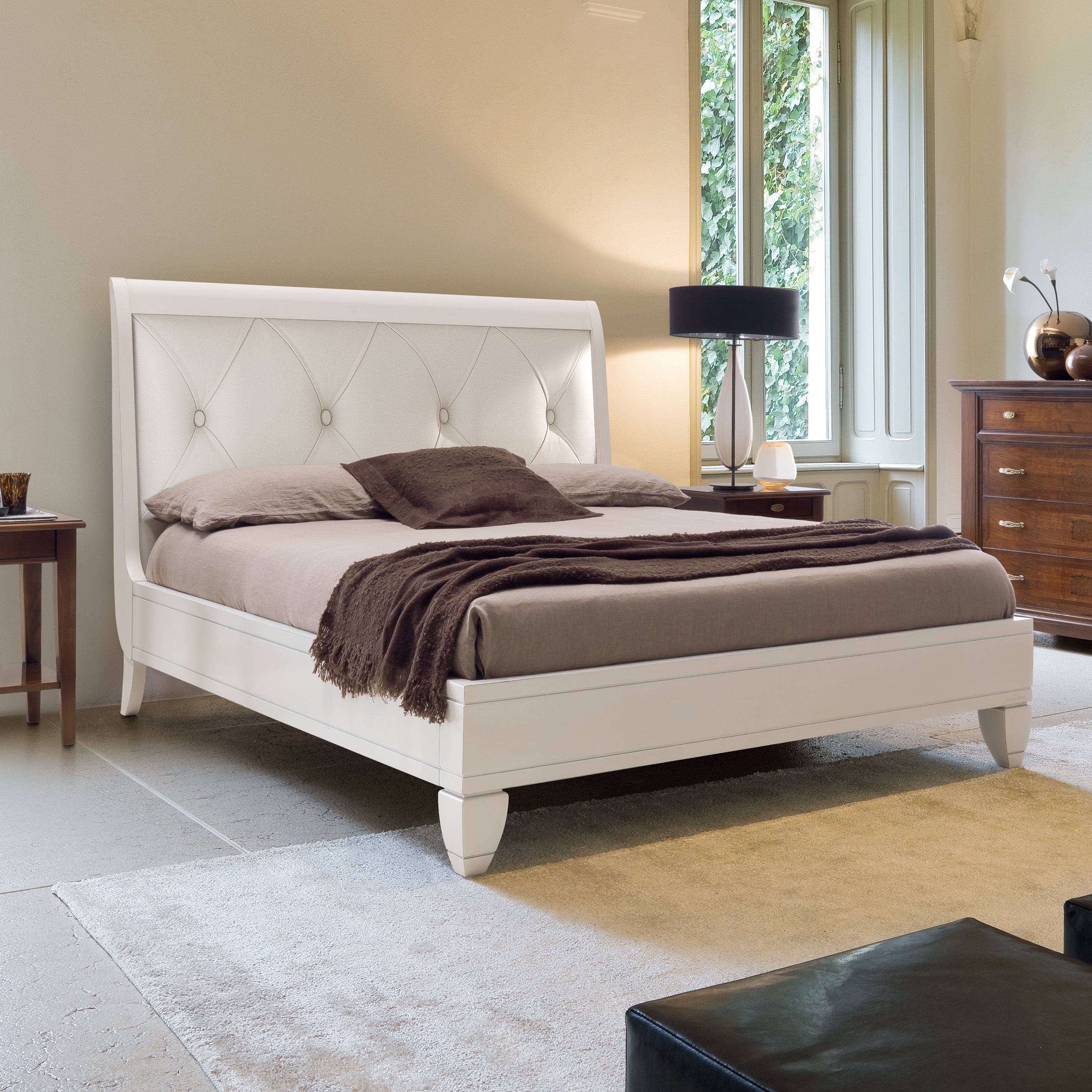 Letto matrimoniale classico in legno di ciliegio laccato bianco, impreziosito da una testata imbottita in vera pelle con lavorazione capitonné. Il contesto raffinato e luminoso è completato da comodini e comò in finitura ciliegio naturale.
