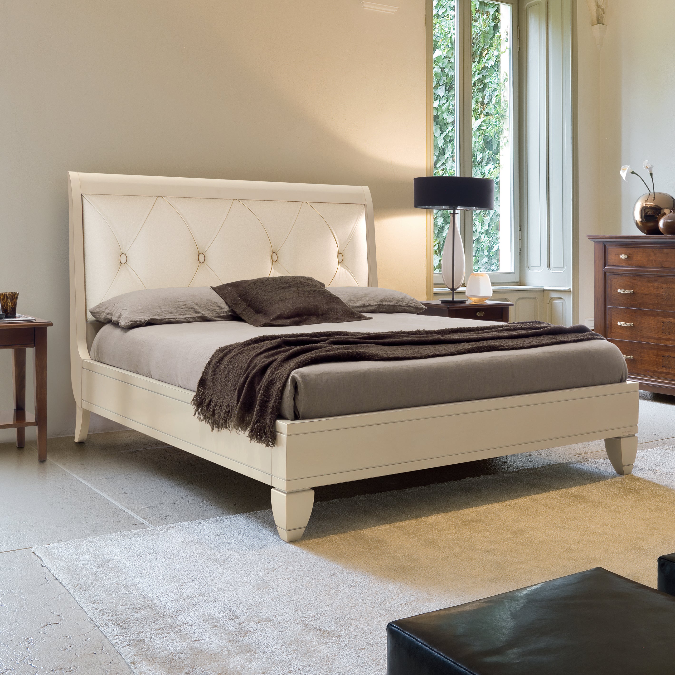 Letto matrimoniale classico in legno di ciliegio laccato color burro, con testata in vera pelle color avorio e lavorazione capitonné. L’ambiente elegante unisce arredi in finitura ciliegio e laccata, con giochi di luce naturale provenienti dalle grandi finestre.