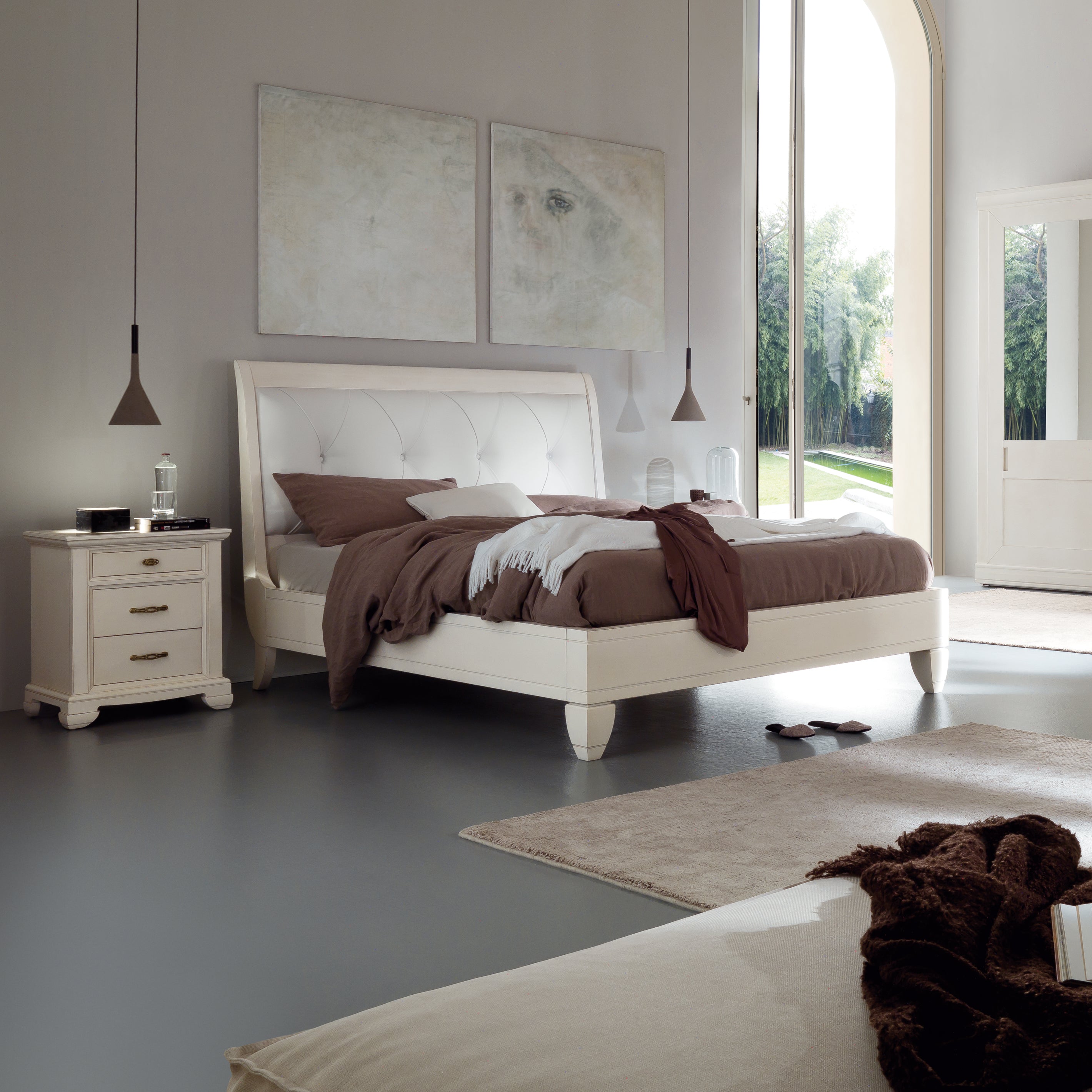 Camera da letto elegante con letto matrimoniale classico in legno di ciliegio laccato color burro, dotato di testata imbottita in vera pelle con lavorazione capitonné. Accanto al letto, un comodino coordinato con tre cassetti e maniglie in ottone. L’ambiente moderno e raffinato è valorizzato da ampie vetrate, pareti chiare e opere d’arte contemporanea.