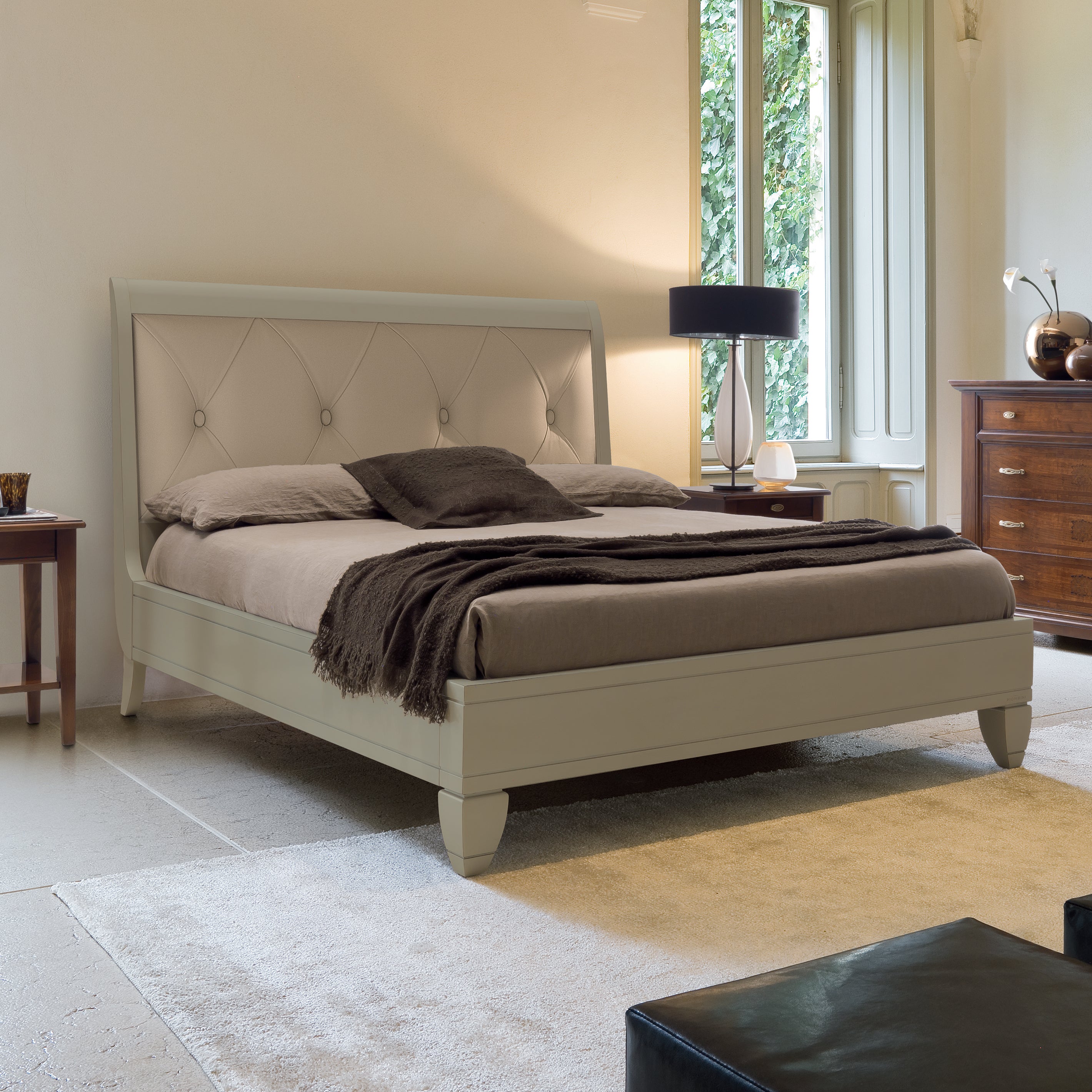 Letto matrimoniale classico in legno di ciliegio laccato color tortora, con testata imbottita in vera pelle e lavorazione capitonné. Il design elegante è accompagnato da comodini e comò in legno ciliegio naturale. La stanza è luminosa, con ampie finestre e arredi raffinati.