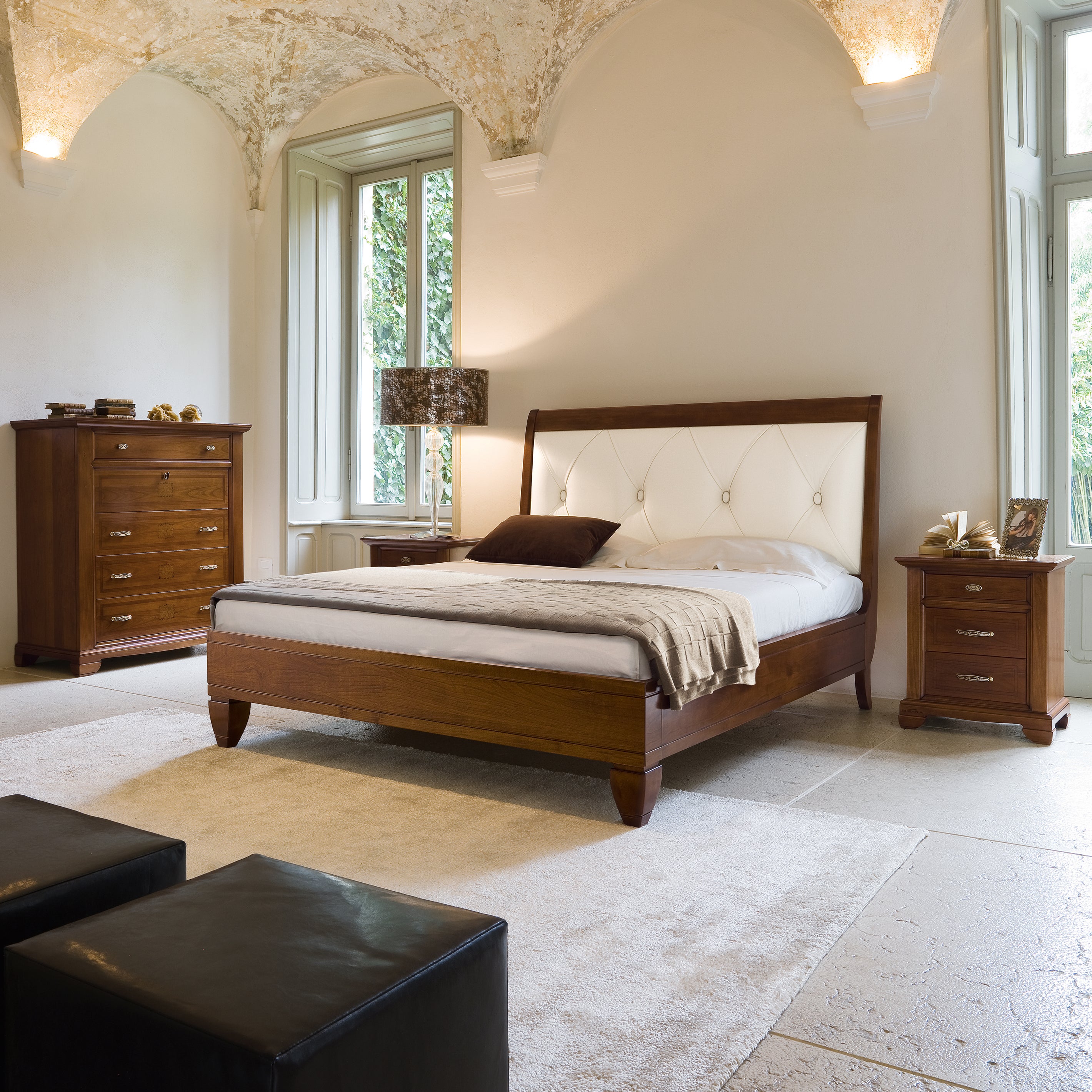 Camera da letto arredata in stile classico con letto matrimoniale in legno di ciliegio e testata imbottita in vera pelle con lavorazione capitonné. Accanto al letto, due comodini coordinati con maniglie in ottone e, sullo sfondo, un comò a quattro cassetti con intarsi decorativi. L’ambiente è raffinato, con soffitto a volta affrescato e grandi finestre che illuminano la stanza.
