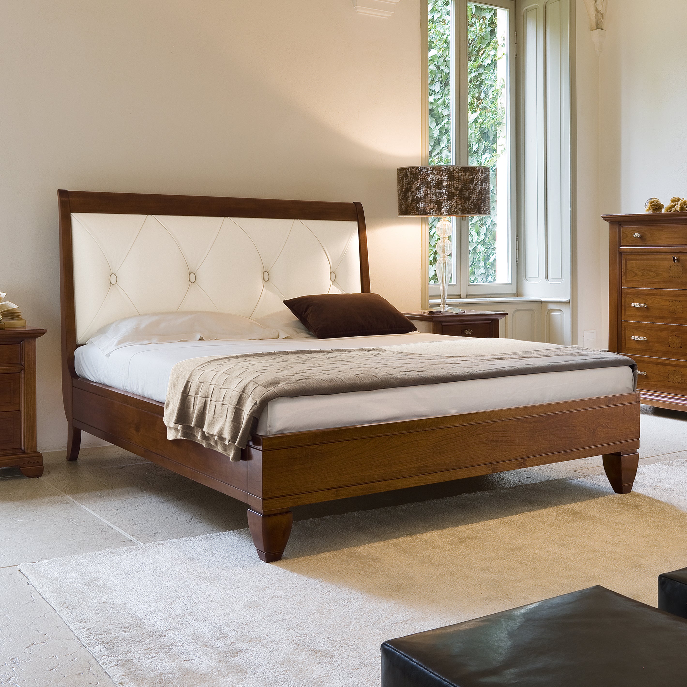 Letto matrimoniale classico in legno di ciliegio con testata imbottita in vera pelle color ecrù, decorata con raffinata lavorazione capitonné. Il design elegante è accompagnato da comodini coordinati e da un comò con intarsi, tutti parte della collezione Arte Piombini. La stanza, luminosa e accogliente, è caratterizzata da ampie finestre e arredi in stile tradizionale.