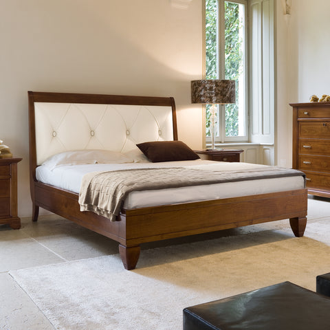 Letto matrimoniale classico in legno di ciliegio con testata imbottita in vera pelle color ecrù, decorata con raffinata lavorazione capitonné. Il design elegante è accompagnato da comodini coordinati e da un comò con intarsi, tutti parte della collezione Arte Piombini. La stanza, luminosa e accogliente, è caratterizzata da ampie finestre e arredi in stile tradizionale.