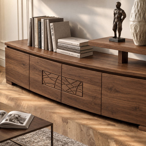 La base in noce della parete Piombini si distende lungo la parete chiara, con frontali dalle venature avvolgenti e un decoro geometrico al centro. Un piano rialzato sostiene una piccola scultura e un vaso. Libri, parquet a spina e un tappeto materico compongono un soggiorno caldo, intimo, vissuto con eleganza.