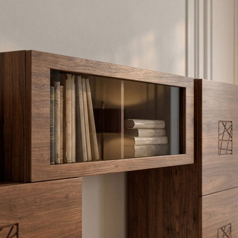 Una cornice ampia in noce racchiude il vetro del pensile Piombini. Al suo interno, libri chiari sono disposti in verticale e in piccole pile. Riflessi ambrati velano la trasparenza, mentre le venature del legno e il vicino frontale inciso creano un equilibrio quieto tra materia, libri e ricordi.