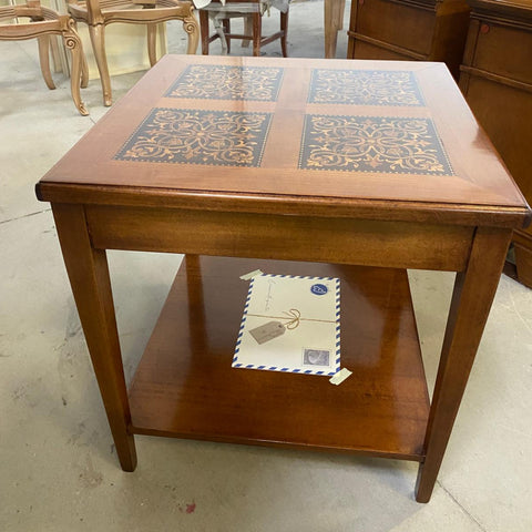 Vista frontale del tavolino quadrato classico Tiziano nella falegnameria. Il piano color ciliegio, decorato con quattro raffinati intarsi floreali su fondo scuro, poggia su quattro gambe dritte e slanciate. Un ampio ripiano inferiore completa il mobile. Le calde tonalità del legno e le linee essenziali trasmettono una sensazione di armonia, solidità ed eleganza discreta.