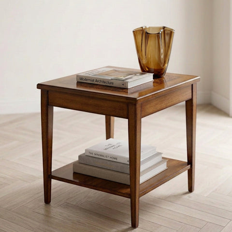 Il tavolino quadrato classico Tiziano, dalle calde tonalità del legno e dalle gambe slanciate, è protagonista di un ambiente luminoso ed essenziale. Sul piano riposano due libri e un vaso in vetro ambrato, mentre il ripiano inferiore accoglie altri volumi. Il pavimento chiaro e le pareti neutre esaltano le proporzioni del mobile, evocando un'atmosfera di quiete, eleganza e piacere quotidiano.