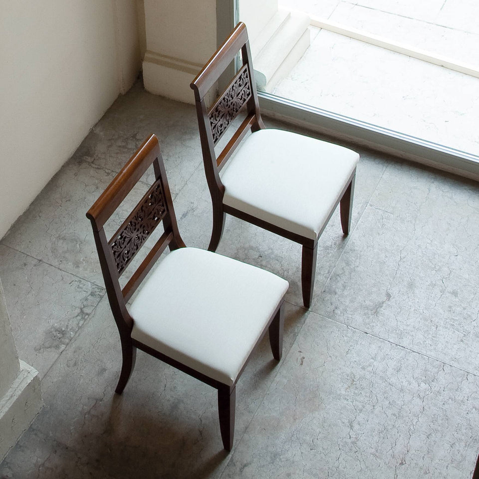 Due sedie classiche Arte, viste dall'alto, mostrano la delicatezza degli schienali in legno finemente traforato e le sedute imbottite color avorio, disponibili in pelle o tessuto. Accanto a una grande vetrata, la luce naturale accarezza le forme e il pavimento in pietra chiara, creando un'atmosfera di quiete ed eleganza discreta.