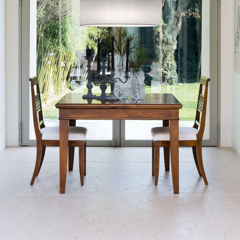 Due sedie classiche Arte in legno, con schienali decorati e sedute chiare disponibili in pelle o tessuto, si fronteggiano intorno a un tavolo. La luce attraversa le grandi vetrate affacciate sul giardino, illuminando il legno e creando un'atmosfera di quiete, armonia e piacere di vivere la casa.