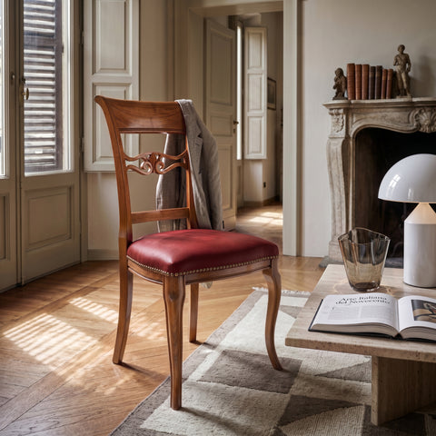 Sedia classica Vittoria in legno massello con seduta imbottita rivestita in vera pelle Siviglia Top Rubino, inserita in un elegante soggiorno italiano illuminato dalla luce naturale con atmosfera calda e ricercata