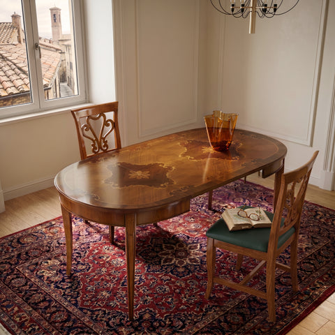 Tavolo ovalino classico allungabile completamente aperto con elegante piano intarsiato in ciliegio, noce e acero, inserito in una luminosa sala da pranzo. La luce naturale esalta le venature del legno, mentre il tappeto classico, le sedie coordinate e l'atmosfera raccolta trasmettono il piacere di accogliere famiglia e ospiti attorno a un tavolo artigianale italiano.