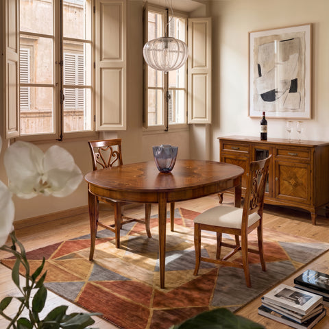 Tavolo ovalino classico allungabile con piano intarsiato in ciliegio inserito in un elegante soggiorno con credenza coordinata, grandi finestre che illuminano l'ambiente con luce naturale, sedie in legno, tappeto dai toni caldi e dettagli che trasmettono un'atmosfera raffinata, accogliente e autenticamente italiana, pensata per vivere la convivialità ogni giorno.