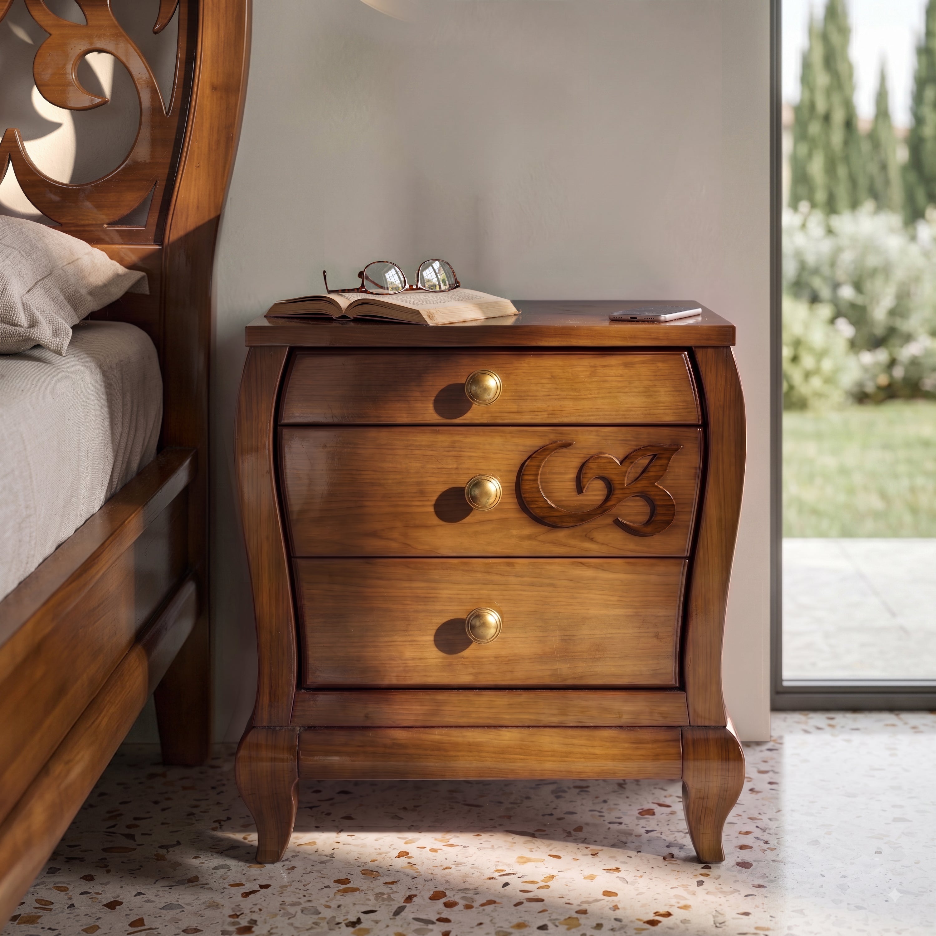 Comodino classico Visconti Piombini in cherry wood with three drawers and central inlay, brass handles, next to a carved bed headboard. On the nightstand are glasses, a book, and a smartphone.