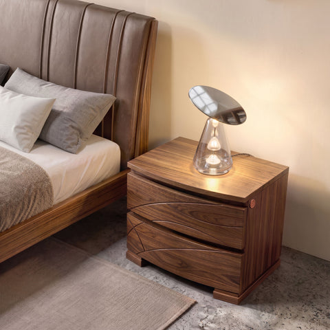 Moderner Nachttisch aus Nussbaumholz, Modell Modigliani, SKU 8271D, mit zwei Schubladen mit wellenförmigen Gravuren und natürlicher Maserung, neben einem Bett mit braunem Lederkopfteil und einer Designlampe.